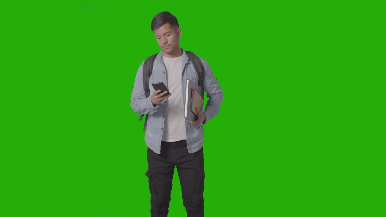 Three Quarter Length Shot Of Male University Or College Student Looking At Mobile Phone Against Green Screen 