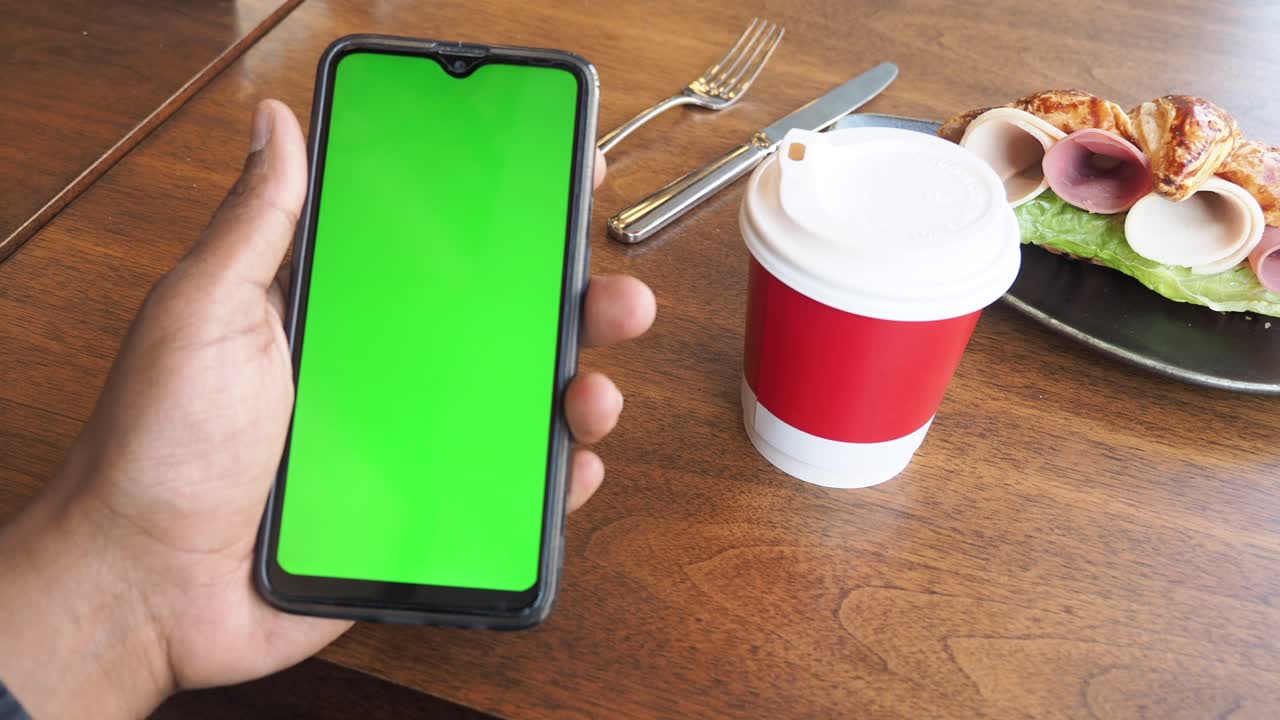 persona con un teléfono de pantalla verde, una taza de café y un sándwich de croissant