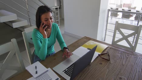 Afroamerikanische-Frau,-Die-Während-Der-Arbeit-Von-Zu-Hause-Aus-Auf-Dem-Smartphone-Spricht-Und-Einen-Laptop-Benutzt