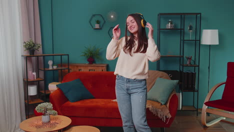 overjoyed young woman in wireless headphones dancing, singing on cozy couch in living room at home