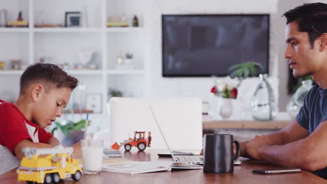 Hispanic-father-and-son-working-opposite-at-dining-room-table,-side-view,-panning,-right-to-left