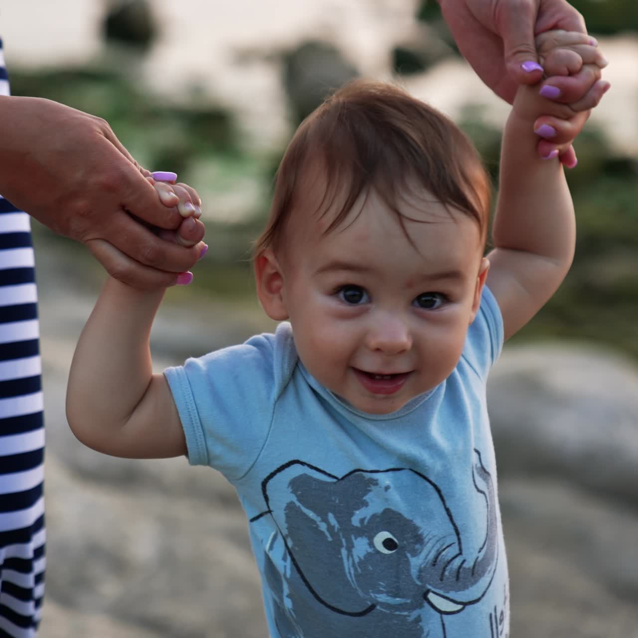Beautiful Caucasian child walking smiling happily. Mother's hands supporting her baby boy. Nature backdrop in blur