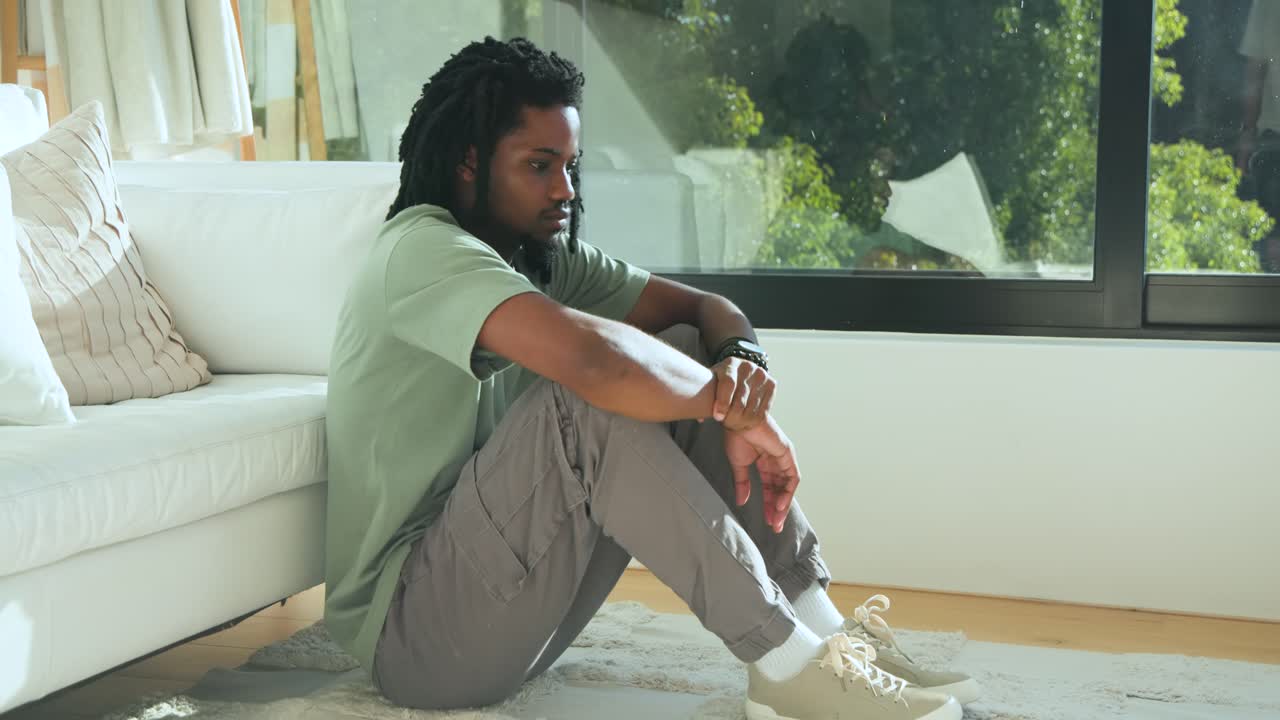 African American man sitting on floor next to white sofa, shifting subtly while window brightening