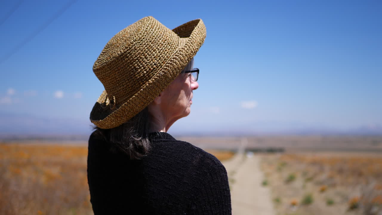 An old woman in summer heat pointing down a dirt road hitchhiking in an empty field of orange California poppy flowers SLOW MOTION