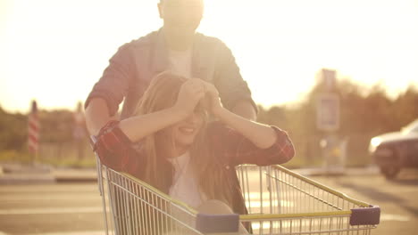 Side-view-of-a-young-man-and-woman-having-fun-outdoors-on-shopping-trolleys.-Multiethnic-young-people-racing-on-shopping-carts.-On-the-parking-zone-with-their