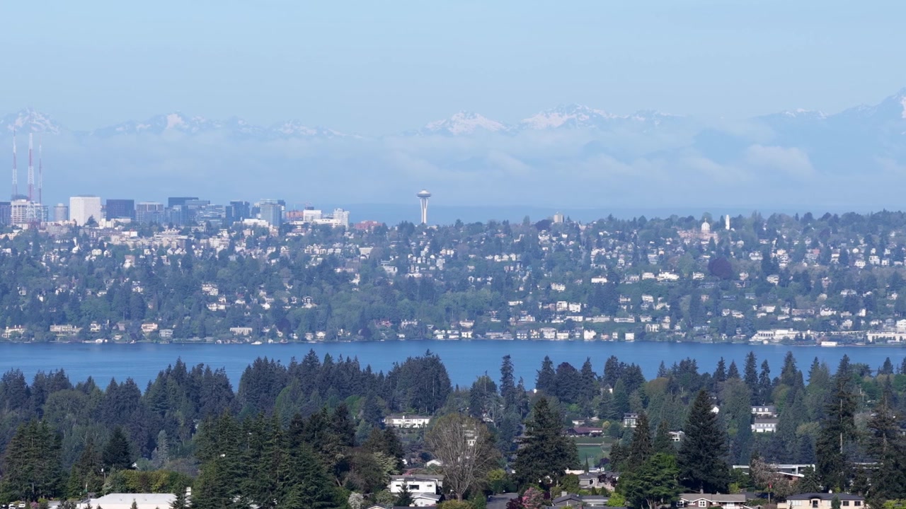 Skyline di Seattle con la lontana Space Needle oltre il Lago Washington, le Montagne Olimpiche innevate, quartieri residenziali con lussureggianti foreste di pini e acque blu calme sotto un cielo limpido e velato. Panorama aereo.