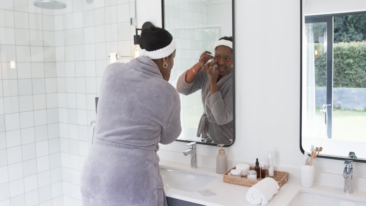 African American woman doing morning facial routine in bathroom applying serum and patches for glow