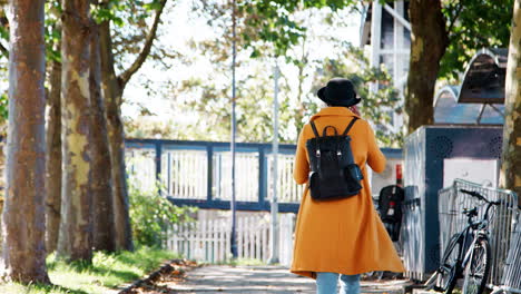 back view of a fashionable young woman wearing a black hat and a yellow coat walking along a treelined street carrying a rucksack on sunny day, close up