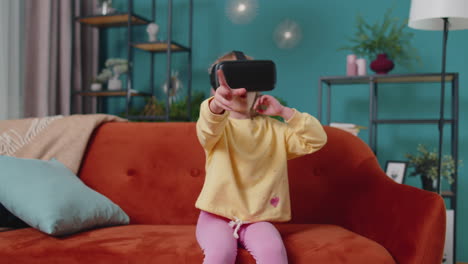 Toddler-girl-sitting-on-home-sofa-using-virtual-reality-headset-helmet-app-to-play-simulation-game