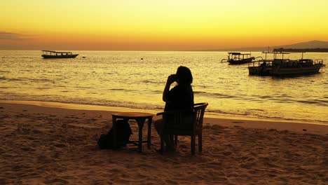 Silhouette-of-girl-talking-on-smartphone,-sitting-on-exotic-beach,-watching-beautiful-sunset-with-yellow-sky-over-anchored-boats,-Bali