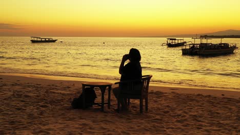 silhouette of girl talking on smartphone, sitting on exotic beach, watching beautiful sunset with yellow sky over anchored boats, bali