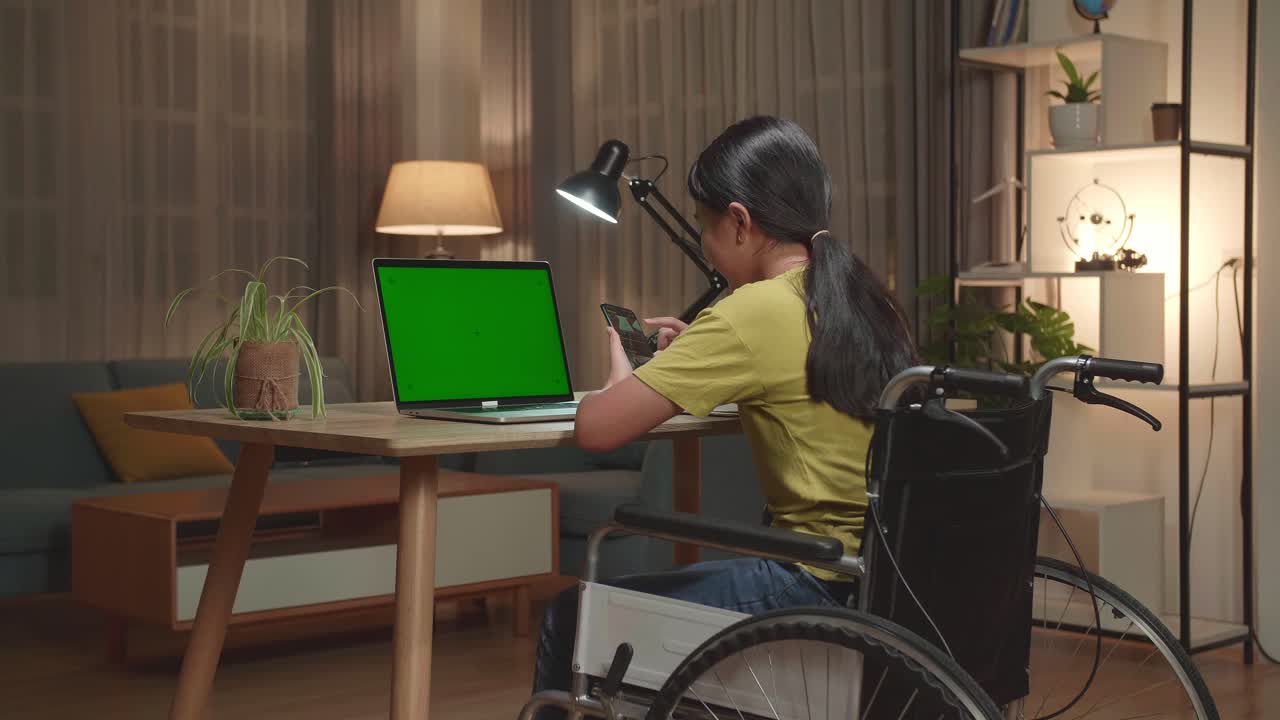 Back View Of Asian Kid Girl Sitting In A Wheelchair While Using Laptop Computer With Mock Up Green Screen And Use Mobile Phone At Home