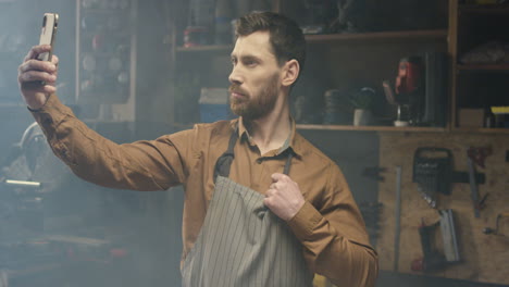 young caucasian male carpenter in apron holding smartphone and making a selfie with mobile phone