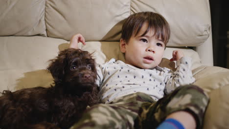 a kid of two years lies on the couch next to the puppy, watching tv