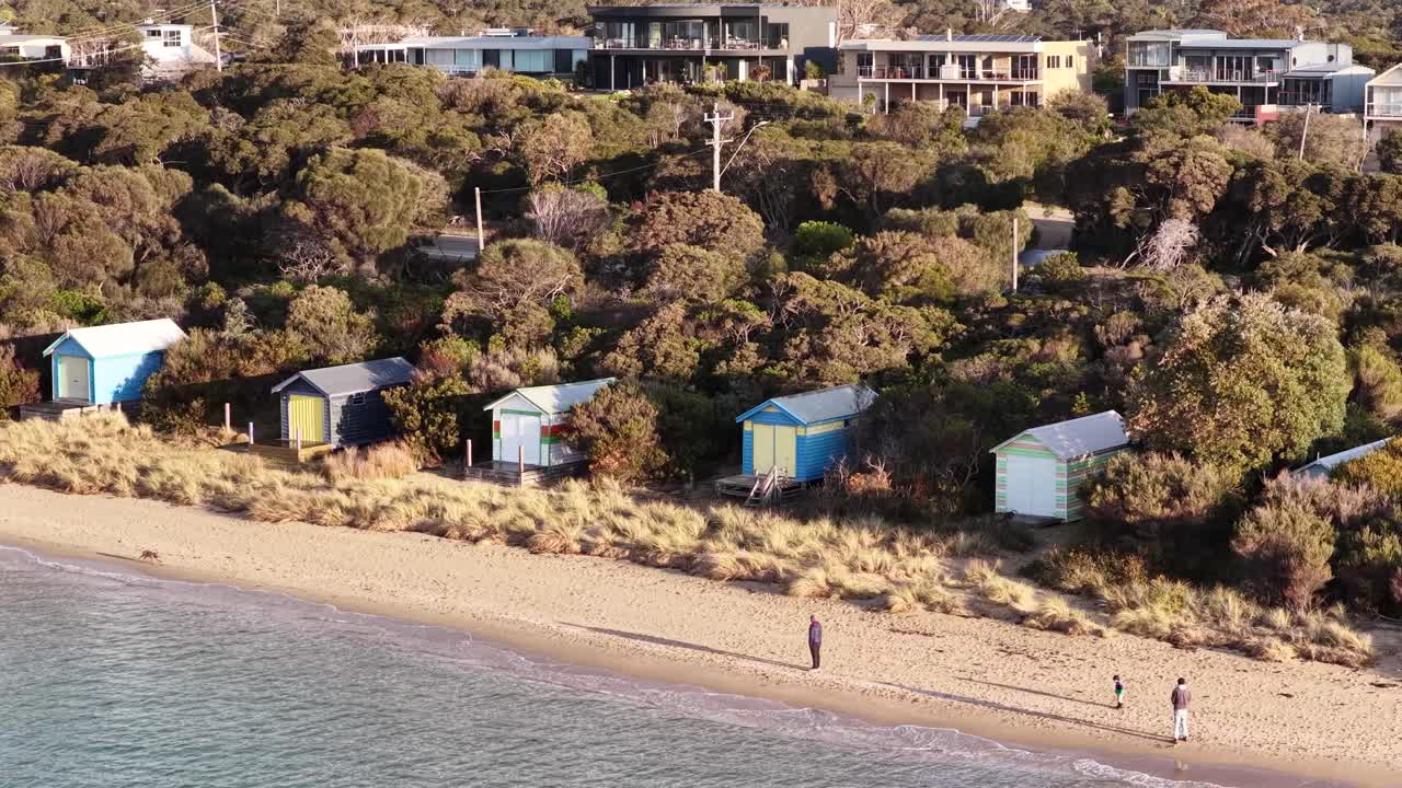 Drone pans above colorful beach huts, sandy shoreline, and coastal vegetation in warm evening light
