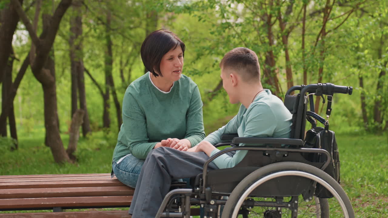 Caregiver Speaking With Young Man In Wheelchair On Park Bench, Engaged Supportive Counseling And Listening In Leafy Environment, Gentle Gestures And Attentive Rapport Under Spring Canopy