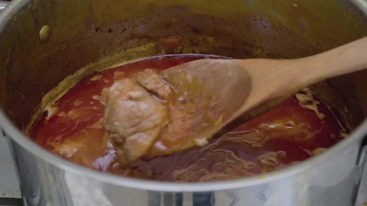 Dettaglio ravvicinato di pulled pork che cuoce in una pentola, bollendo e venendo mescolato con un cucchiaio di legno. La salsa ricca e rossastra bolle e sobbolle, creando un pasto delizioso