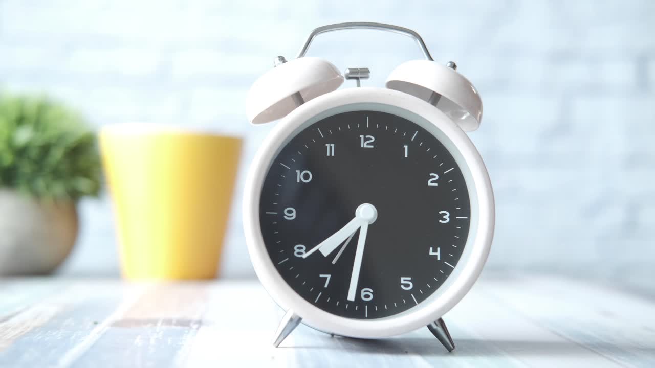White alarm clock on a table with a yellow cup and plant