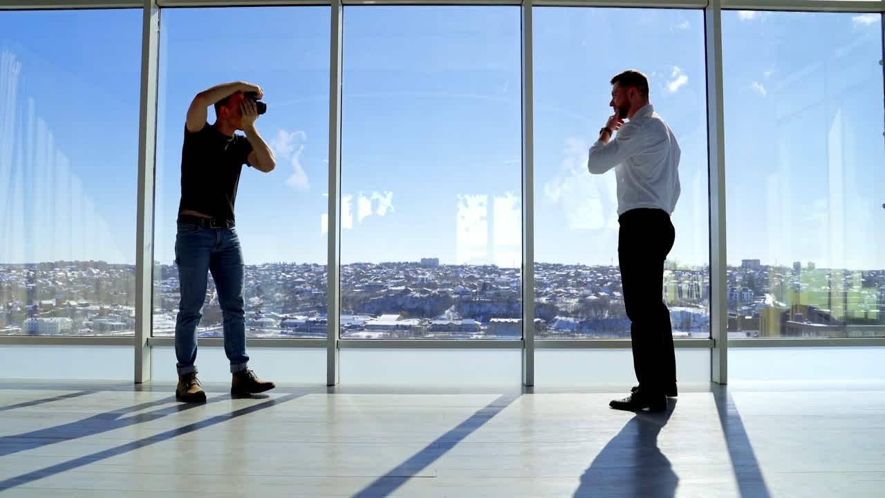 Man photographing businessman in light studio. Handsome man in white shirt and trousers posing on camera next to large windows in office in a bright day. Profile view.