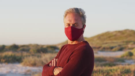 Portrait-of-happy-caucasian-man-in-face-mask-standing-with-arms-crossed-on-sunny-day-at-the-beach