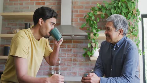 biracial young man drinking coffee while talking with father in kitchen at home