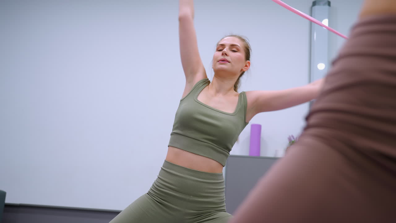 primer plano de una dama con una camiseta marrón participando en ejercicios de fitness estirándose con una cinta en un gimnasio, otra dama con un traje verde fluido cerca, equipos dispuestos en una mesa