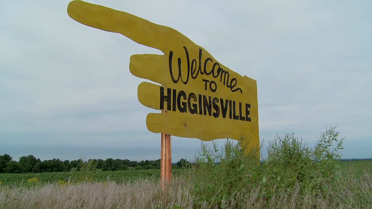 un gran cartel amarillo apunta a higginsville missouri desde un campo agrícola