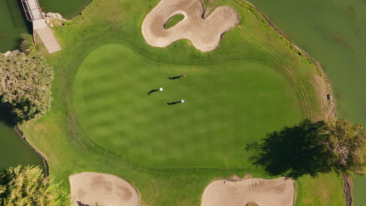Veduta aerea diretta dall'alto con drone di tre golfisti che imbucano e camminano su un green circondato dall'acqua.