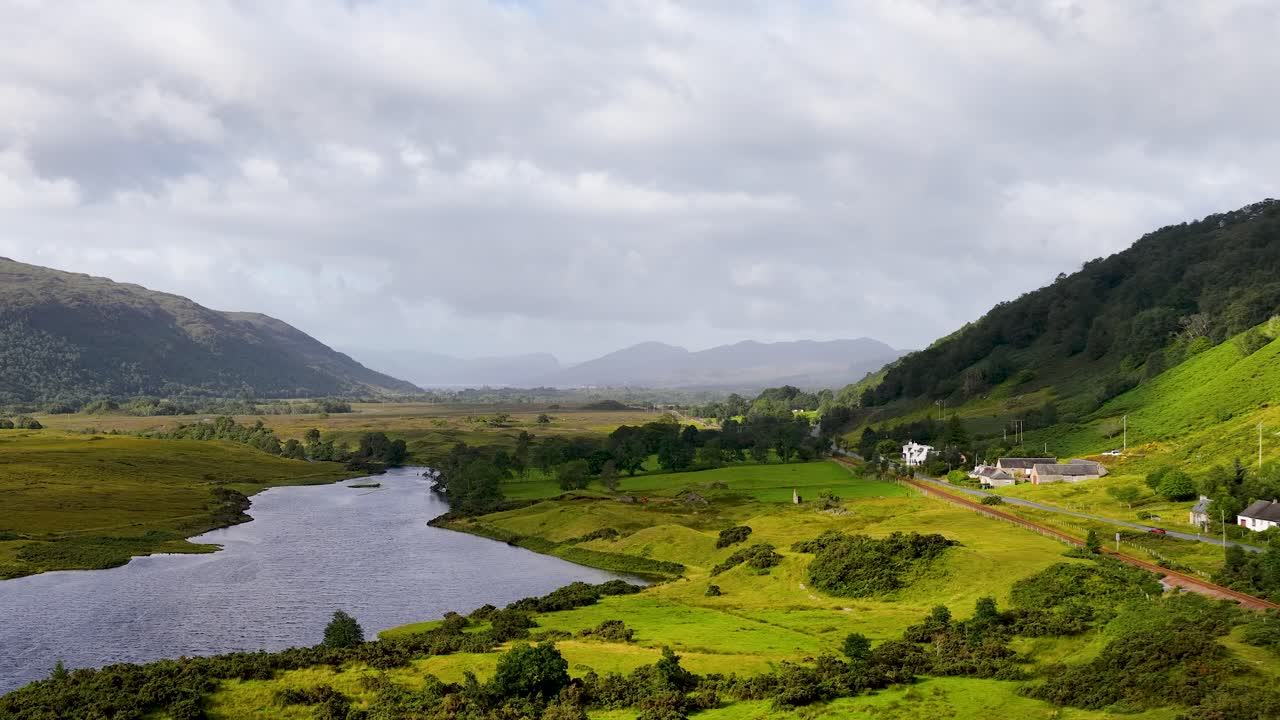 Aerial camera glides above serene lake, lush green valley, and distant mountains under soft daylight