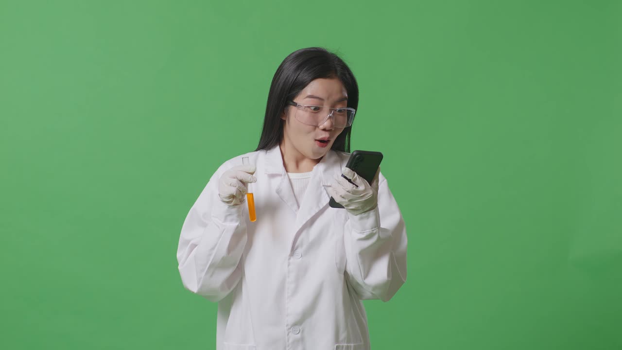실험관 안의 오렌지색 액체와 함께 스마트폰을 보고 실험실의 녹색 스크린 배경에 서있는 동안 와우라고 말하는 아시아 여성 과학자