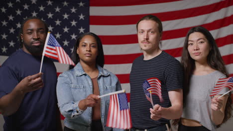 Studio-Portrait-Shot-Of-Multi-Cultural-Group-Of-Friends-Waving-Miniature-Stars-And-Stripes-Flags-In-Front-Of-American-Flag-Celebrating-4th-July-Independence-Day-Party-2