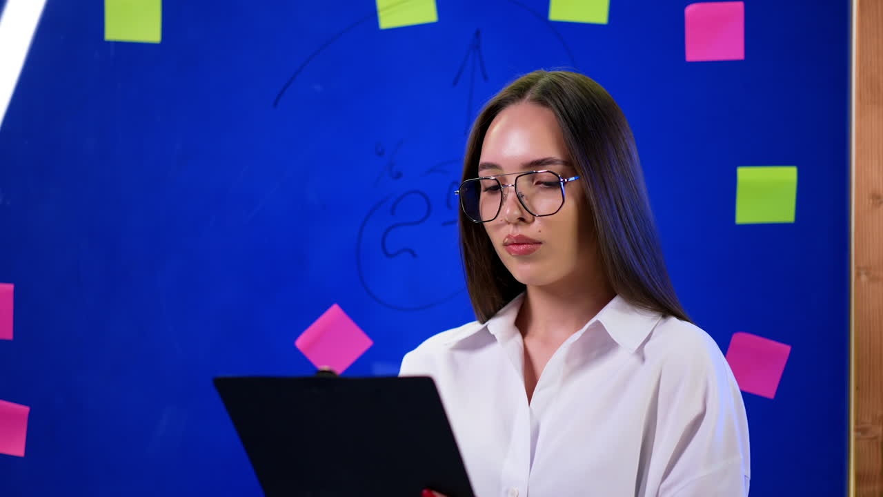 Serious Caucasian woman in glasses looks focused on the clipboard in her hands. Portrait of a modern young business lady near glass board.