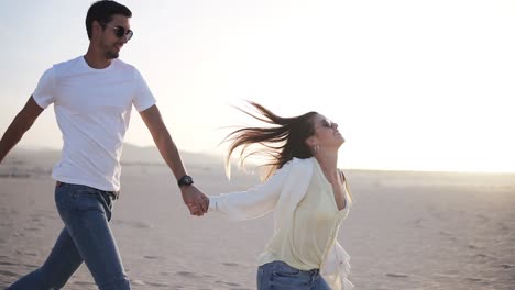 Couple-holding-hands-running-having-fun-under-sun-in-playful-and-romantic-relationship-under-sun-and-blue-sky-in-desert.-Two-young-lovers-cheerful-together-on-romance-in-summer.-Wearing-casual-clothes-and-sunglasses