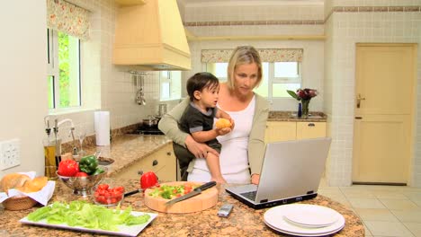 madre joven en la cocina trabajando en una computadora portátil sosteniendo a su bebé