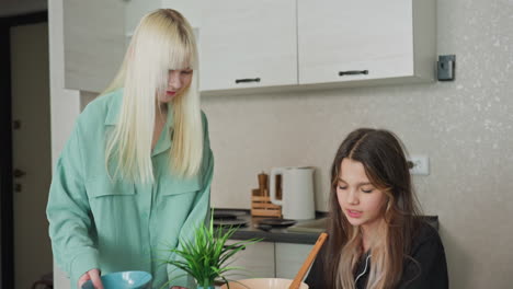 two sisters share playful moment in bright kitchen as seated sibling holds mixing bowl on head while standing sister taps bowl rhythmically with spoon laughter and warm sibling bond