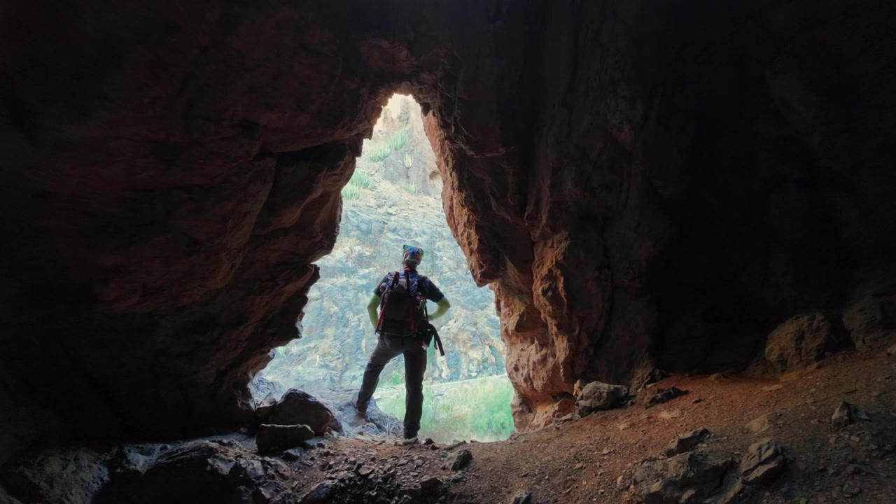 Adventurous man with a backpack standing at the entrance of a dark cave, admiring the sunny landscape of a beautiful ravine in Spain