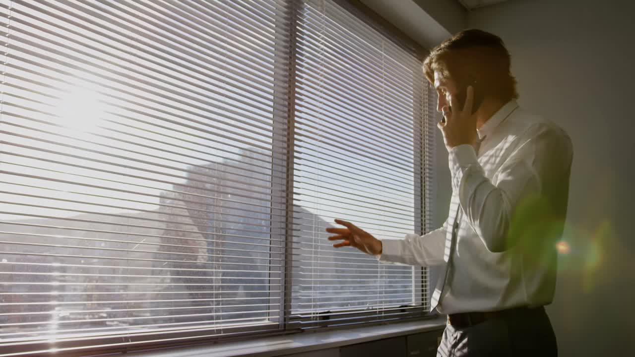 Side view of young Caucasian male executive talking on mobile phone near window in modern office 4k