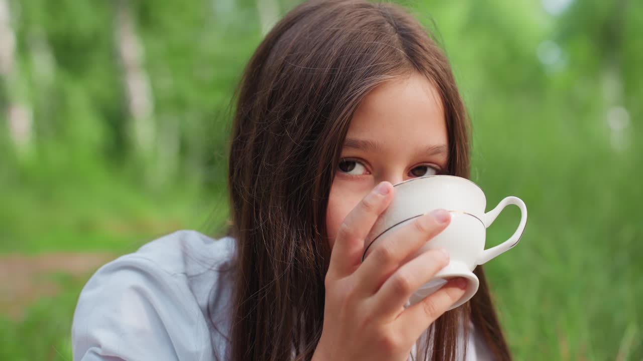 밝은 녹색 나무와 풀로 둘러싸인 야외에서 컵을 들고 음료를 마시는 어린 소녀의 클로즈업 사진이에요. 따뜻한 여름날 부드러운 햇빛을 받으며 평화로운 자연의 순간을 즐기고 있어요.
