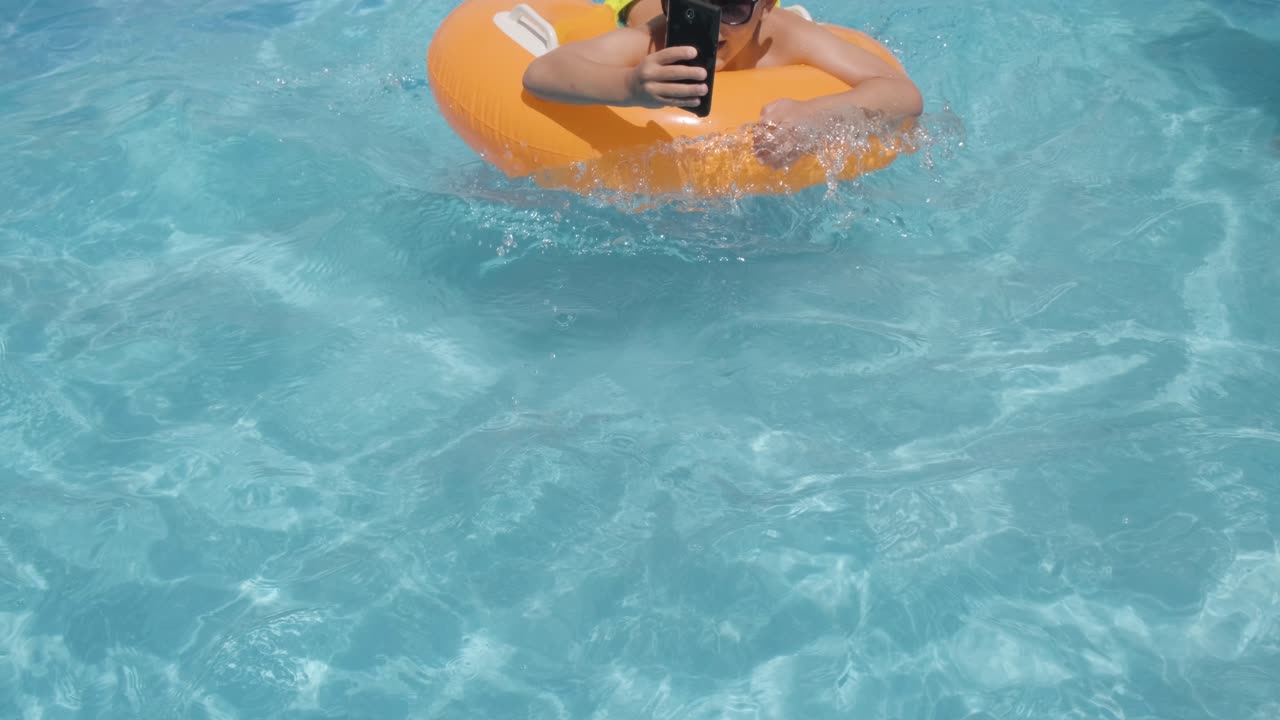 joven con gafas de sol usando un celular en un flotador naranja mientras se relaja en la piscina