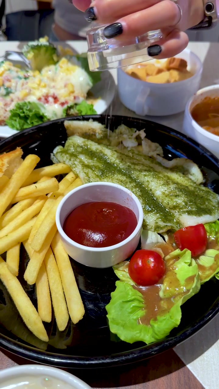 Primo piano di una donna che condisce una bistecca di pesce con patatine fritte, insalata e ketchup in una luminosa illuminazione da ristorante