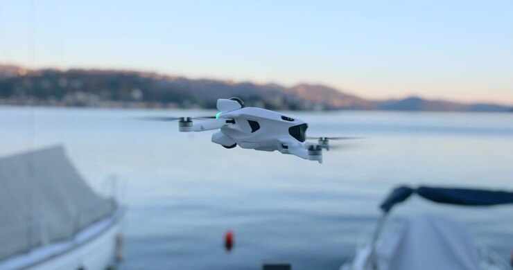 Close up of a compact white drone hovering in mid-air over a lake with boats at sunset