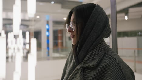 lady wrapped in scarf and glasses turns around while standing in illuminated indoor setting with soft lighting and reflective surfaces in background