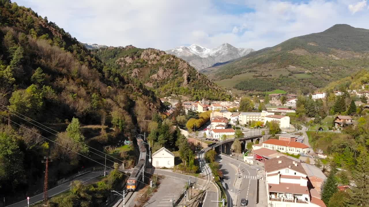 antena: tren regional entrando en una estación en los pirineos con picos nevados al fondo