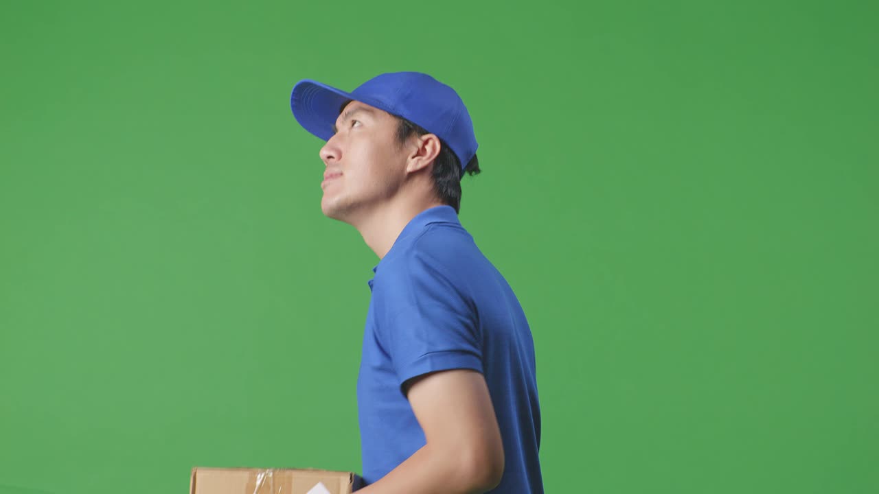 Primo piano laterale di corriere maschio asiatico sorridente in uniforme blu che cammina e si guarda intorno mentre consegna un pacco su sfondo green screen in studio