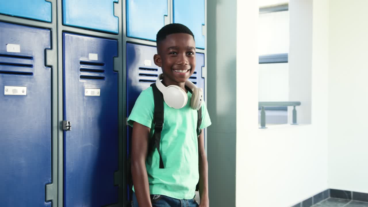 Bambino afroamericano con maglietta color menta e jeans che usa il telefono, che guarda la telecamera in posa accanto agli armadietti