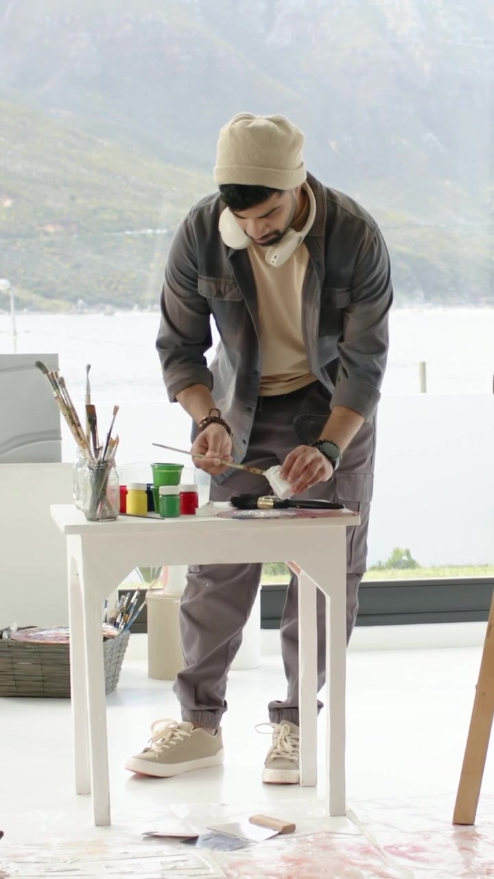 Vertical video: Approaching table, man in beanie mixing paints, prepping palette near lake window