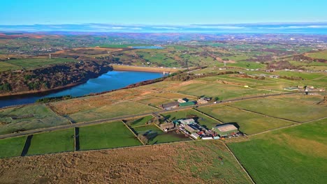 Imágenes-Aéreas-De-Drones-Del-Tráfico-En-Una-Carretera-Rural-Con-La-Autopista-M62-A-Lo-Lejos-En-Agua-Scammonden-Con-Lago-De-Tierras-De-Cultivo,-Embalse-Y-Amplias-Vistas-Abiertas-De-Las-Colinas-Peninas