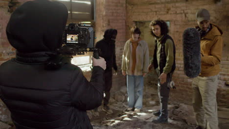 cameraman and team production recording a movie scene with a redheaded girl and man wearing black balaclava in a ruined building 1