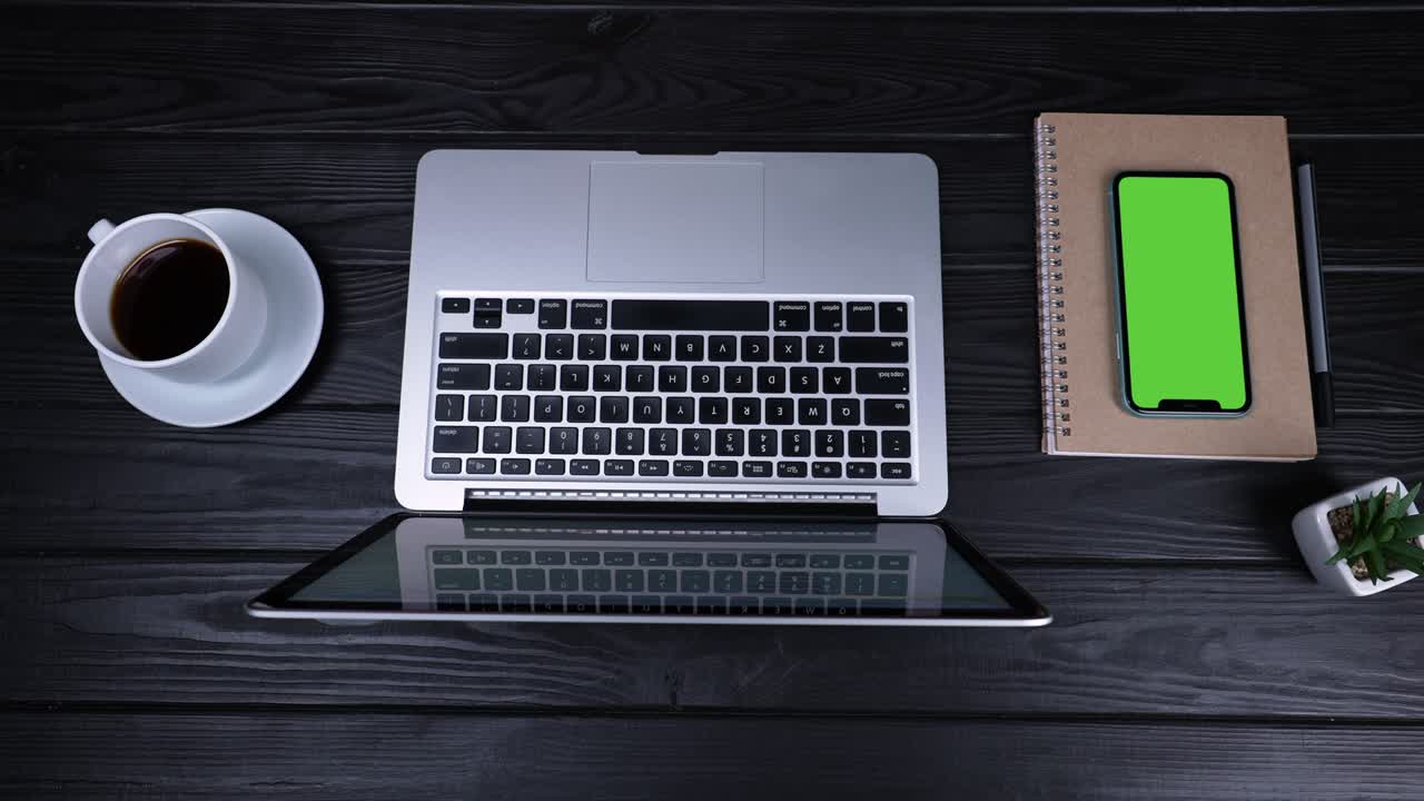 On the table is an open laptop, a cup of coffee, a notepad, a pen and a smartphone with a green screen and a chrome key. Top view of the workplace prepared for work. Slow motion. Close up