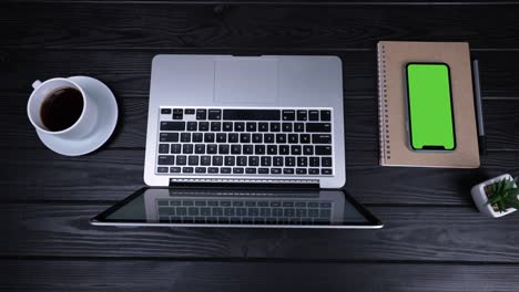 on the table is an open laptop, a cup of coffee, a notepad, a pen and a smartphone with a green screen and a chrome key. top view of the workplace prepared for work. slow motion. close up